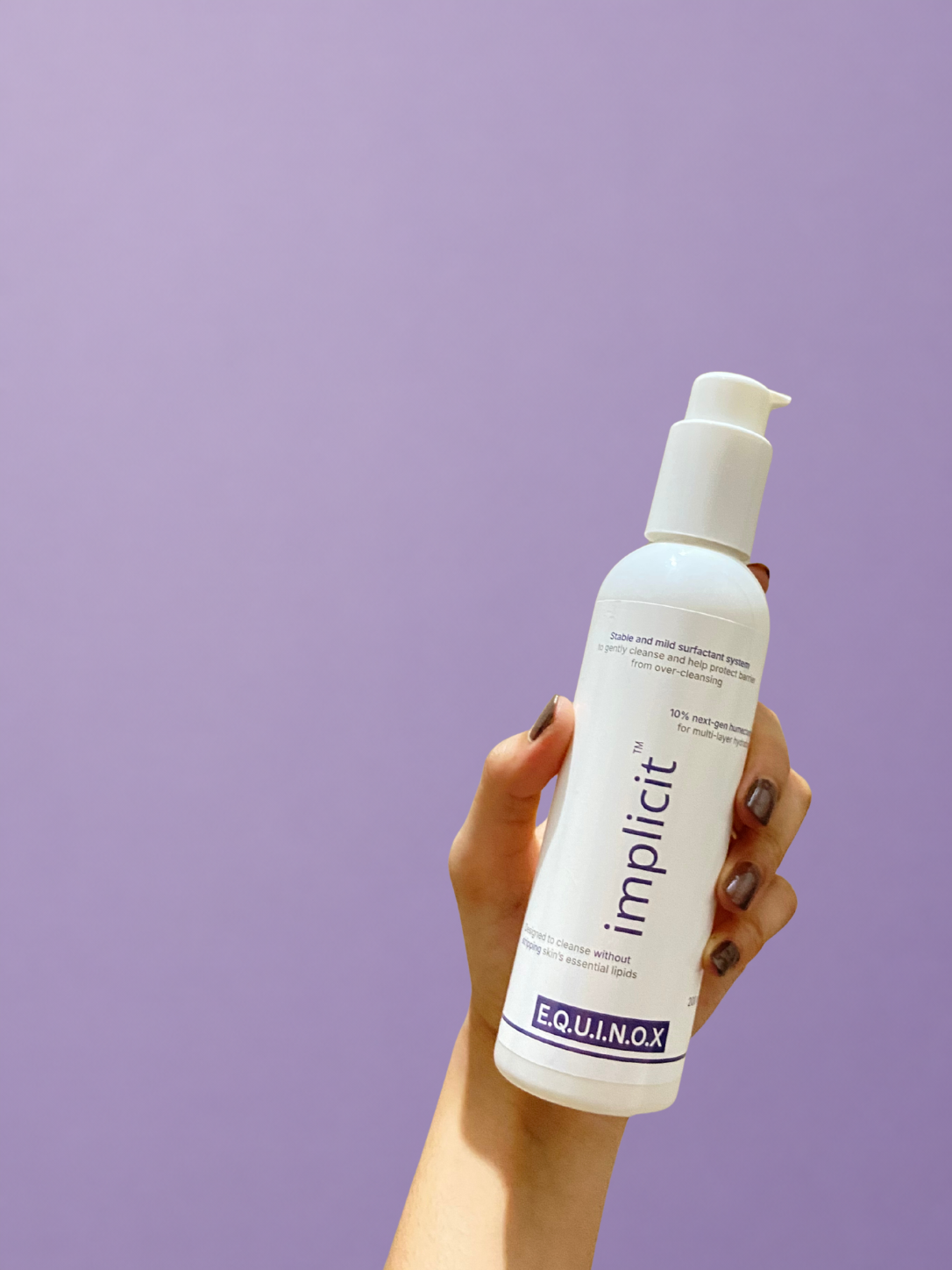 Hand holding an implicit™ E.Q.U.I.N.O.X. gentle face and body cleanser pump bottle against a soft lavender background.